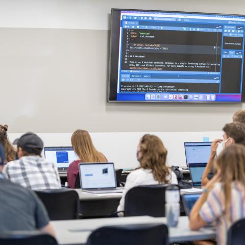 picture of classroom with large tv screen showing code on black background. students in rows looking up at screen