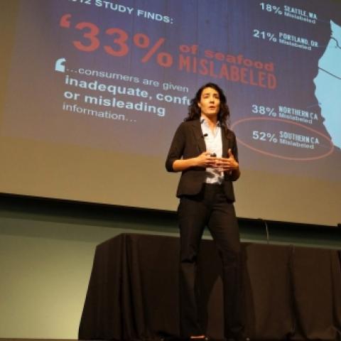 Woman presents on stage with projection on screen behind her
